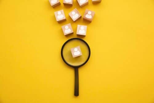 talent acquisition: lente di ingrandimento su sfondo giallo che analizza blocchi di legno con sagoma di persone